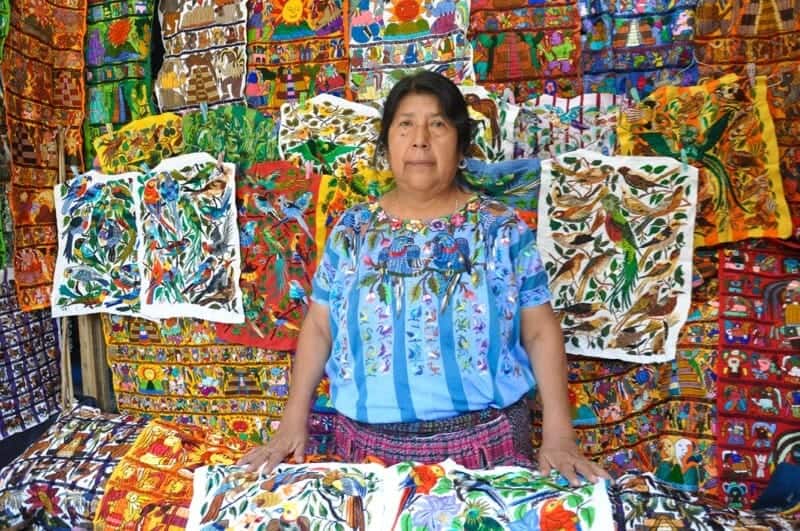 Vendor in blue blouse standing in front of embroidery stall in Chichicastenango in Guatemala.