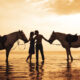 Couple horseback riding on the beach at sunset.