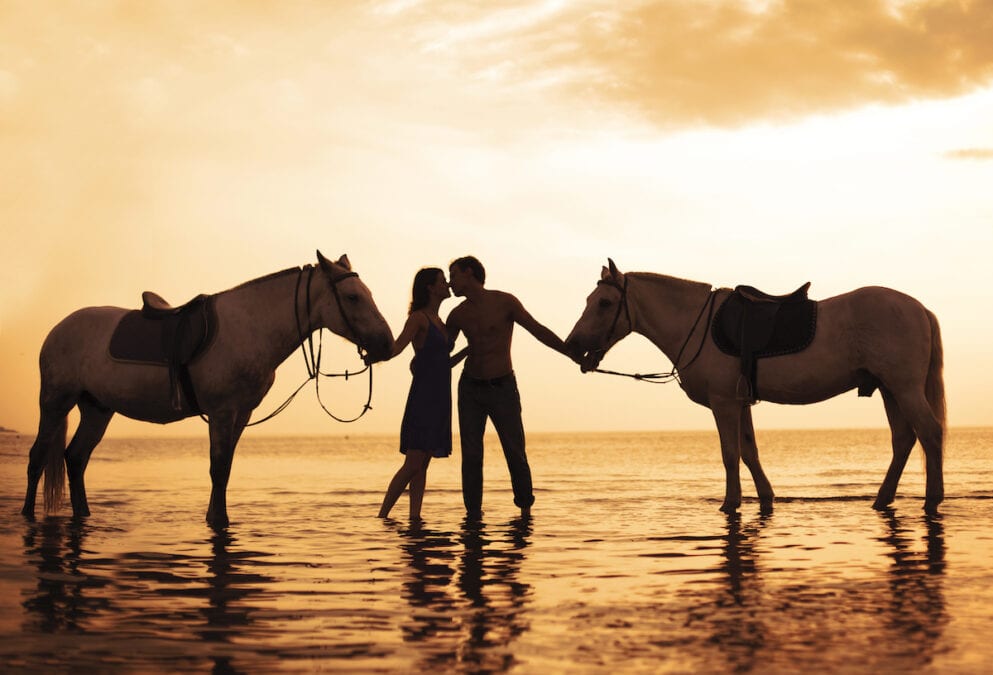 Couple horseback riding on the beach at sunset.