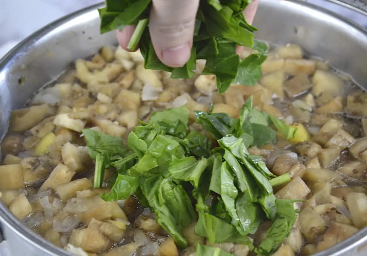 Woman dropping drop handfuls of spinach into a pumpkin and eggplant mixture.