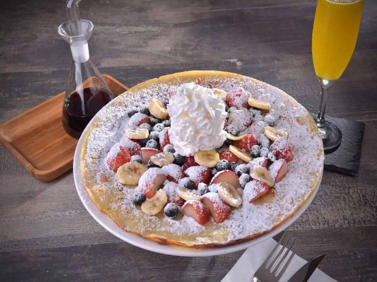 A crepe on a plate covered with fruit, powdered sugar, icing, and whipped cream
