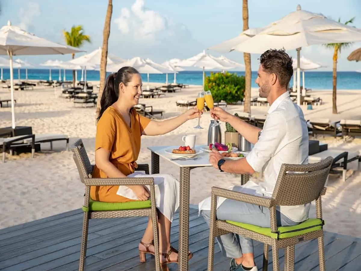 A man and a woman sitting a table outside with drinks and breakfast on the beach