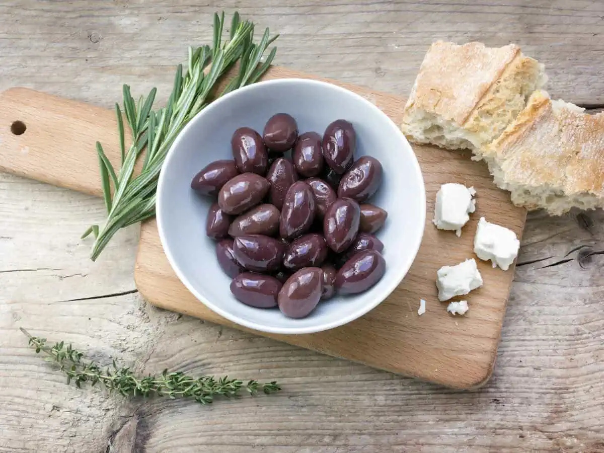 Kalamata black olives in a white bowl, bread, feta cheese and herbs garnish on a rustic wooden table.